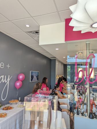 Bubbly pink-and-gray beauty salon interior with stylists in pink shirts tending seated clients, a clear hair-extension display case, snack table with pink balloons, and a glass storefront