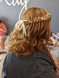 Auburn curly half-up braided crown styled with a pearl-and-crystal hair vine and ivory ribbon bow, salon-ready wedding hairstyle.