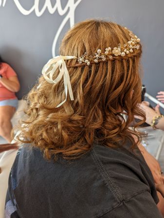 Auburn curly half-up braided crown styled with a pearl-and-crystal hair vine and ivory ribbon bow, salon-ready wedding hairstyle.