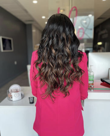 Back view of client with long wavy brunette hair and caramel balayage, wearing a bright pink blazer in a modern hair salon