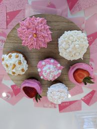 Top-down view of eight pink-and-white decorated cupcakes arranged on a round wooden board — assorted piped frosting styles, edible pearl sprinkles and fresh strawberry halves on a bakery dessert display.