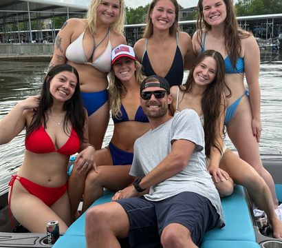 Seven friends in swimsuits smiling and posing on a blue boat at a lake marina, summer scene with docks and a cloudy sky.