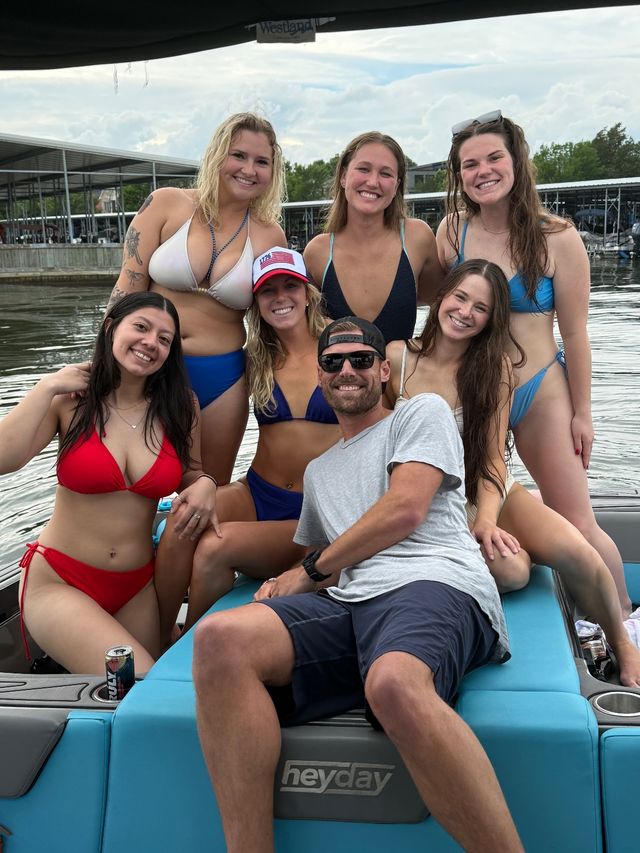 Seven friends in swimsuits smiling and posing on a blue boat at a lake marina, summer scene with docks and a cloudy sky.