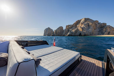 Sunlit luxury yacht deck with cushioned lounge and small American flag, cruising on deep-blue ocean toward rugged rocky coastline and sandy beach.