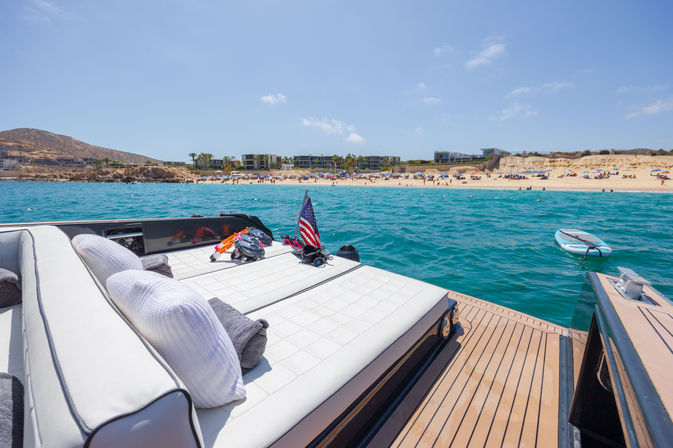 Luxury yacht sunbed with pillows, snorkeling gear and a small American flag anchored off turquoise waters near a sandy beach with a paddleboard, coastal resort buildings and hills under a clear blue sky