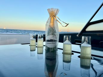 Champagne bottle in a gold-tied gift bag and four bubbly flutes on a glossy yacht table with sailboats on the ocean horizon at sunset