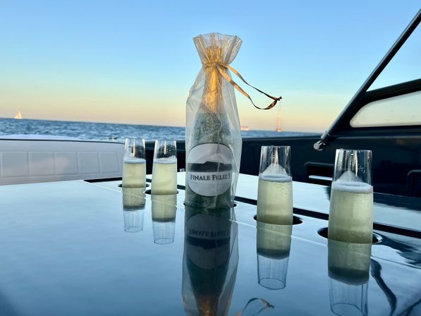 Champagne bottle in a gold-tied gift bag and four bubbly flutes on a glossy yacht table with sailboats on the ocean horizon at sunset