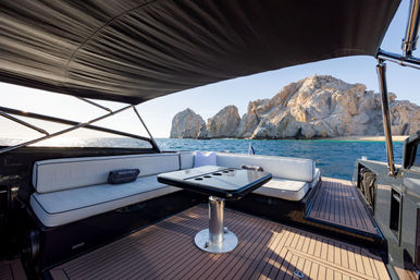 Sunlit luxury yacht lounge with white cushioned seating, central table and black canopy over a teak deck, looking out over turquoise sea and dramatic rocky coastline.