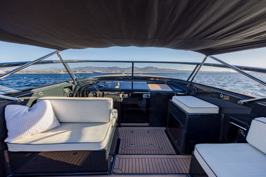 Sunlit luxury motorboat helm with white cushioned benches, teak deck and canvas bimini, overlooking blue ocean and distant coastal mountains.