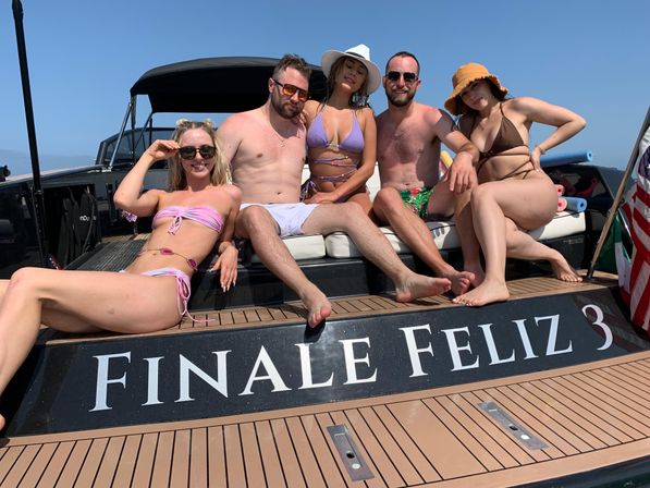 Group of five adults in colorful swimwear lounging on the stern of a motorboat under a clear blue sky, enjoying a sunny summer boat party on open water.