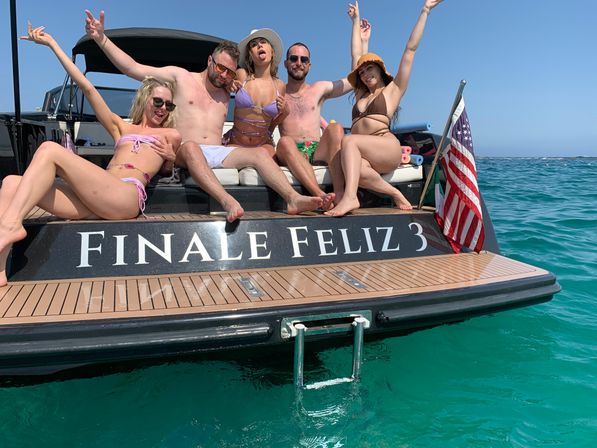 Five friends in colorful swimsuits laughing and posing on the stern of a motorboat with an American flag and clear turquoise ocean on a sunny day.