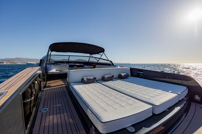 Sun-soaked yacht sun deck with twin cushioned sunbeds, teak flooring and black bimini, sparkling ocean waters and distant coastline under a clear sky