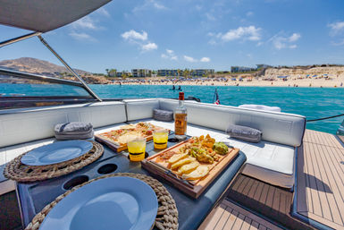 Deckside picnic on a luxury yacht — charcuterie boards, chips, guacamole and cocktails overlooking turquoise water, a sunny sandy beach and coastal villas