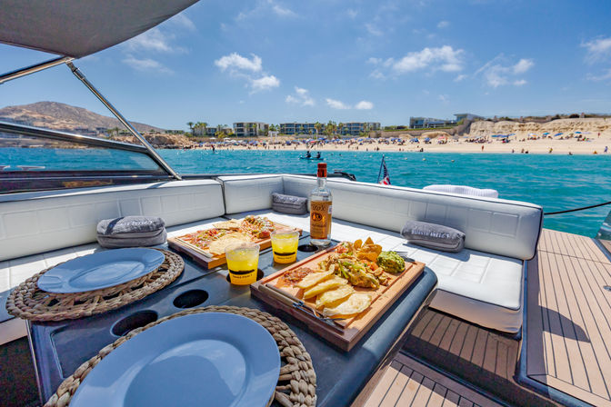 Deckside picnic on a luxury yacht — charcuterie boards, chips, guacamole and cocktails overlooking turquoise water, a sunny sandy beach and coastal villas