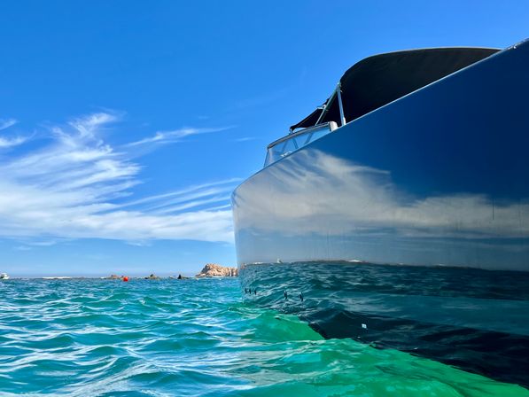 Low-angle coastal view of a sleek motorboat hull reflecting turquoise ocean waves beneath a bright blue sky with wispy clouds and a rocky shoreline in the distance.