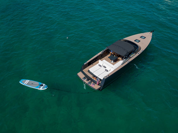 Aerial view of a sleek black-and-teak motorboat anchored in clear turquoise water with cushioned sun pads on deck and a blue-and-white paddleboard tethered alongside.