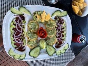 White appetizer platter on outdoor patio with mini tostadas topped with guacamole arranged around a tomato rose, cucumber slices and red onion over sauce in side compartments, with a bowl of tortilla chips and a soda can nearby.