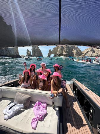 Boat party of friends in pink cowboy hats holding face-cutout props, cruising turquoise waters toward El Arco rock arch at Cabo San Lucas on a sunny day with other boats nearby.