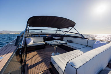 Sunlit luxury boat deck with white cushioned U-shaped seating, central table, teak flooring and black bimini top, cruising on calm blue sea with distant shoreline under a clear sky.