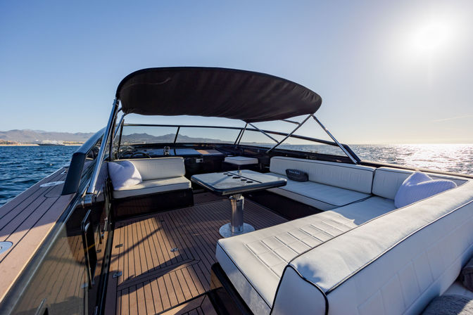 Sunlit luxury boat deck with white cushioned U-shaped seating, central table, teak flooring and black bimini top, cruising on calm blue sea with distant shoreline under a clear sky.