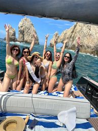 Six women in bikinis cheering on a boat for a bachelorette trip, one wearing a bride sash, posing in front of El Arco rock formation at Cabo San Lucas, Mexico