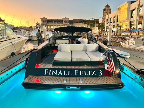 Luxury motorboat docked at a busy marina at sunset, cushioned sunbed on the stern, glowing turquoise underwater lights, American and Mexican flags flying, waterfront shops and palm trees in the background.
