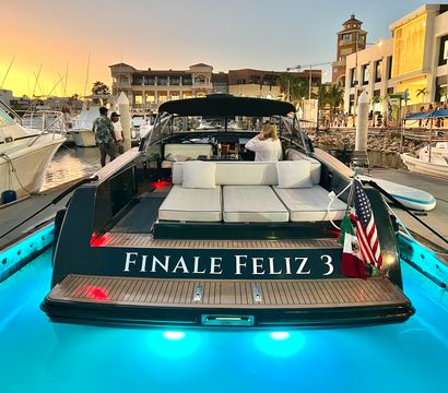 Luxury motorboat docked at a busy marina at sunset, cushioned sunbed on the stern, glowing turquoise underwater lights, American and Mexican flags flying, waterfront shops and palm trees in the background.