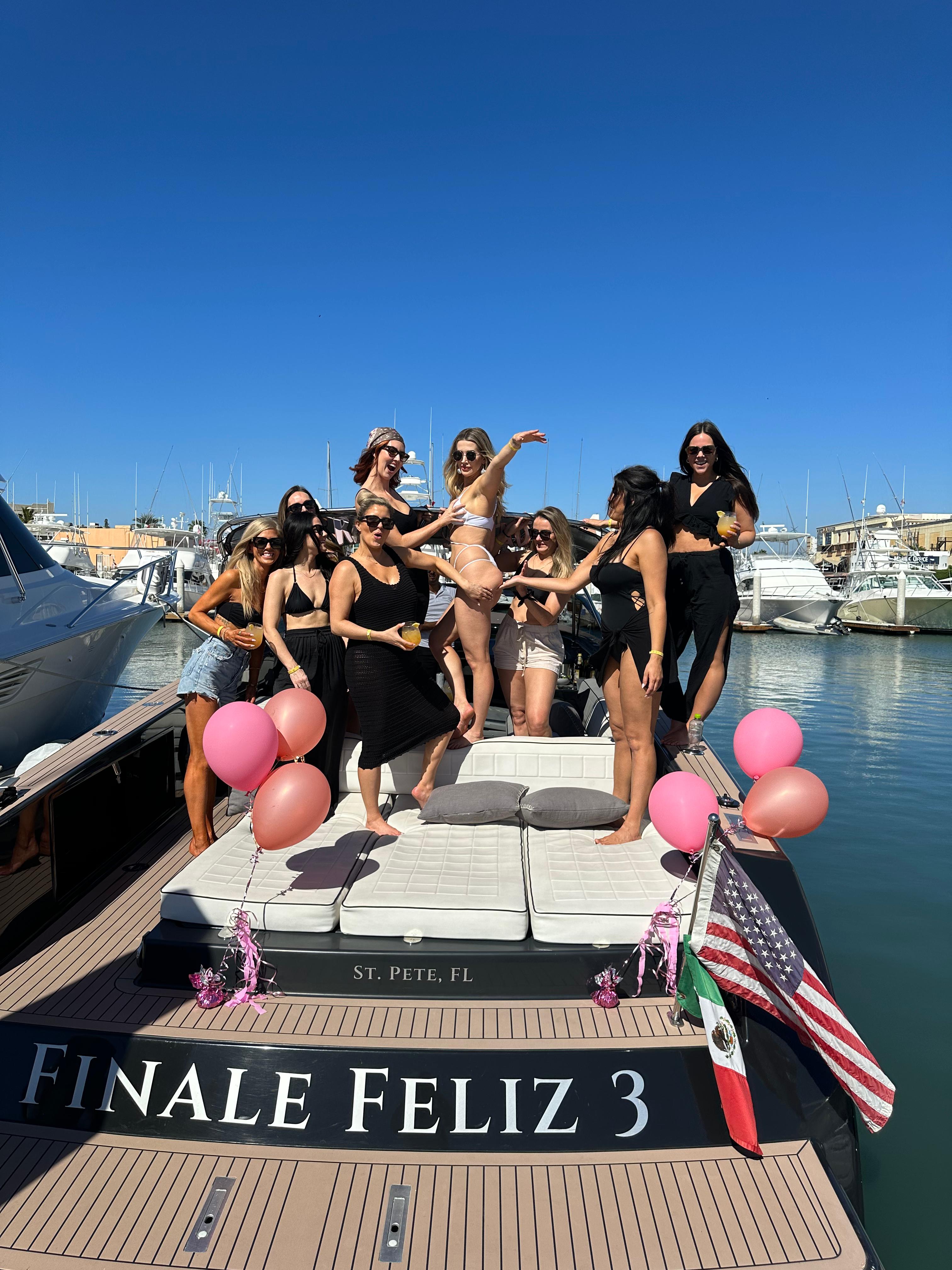 Group of women in swimsuits and summer outfits laughing and posing with drinks and pink balloons on a yacht sun pad at a sunny St. Pete, Florida marina, with blue sky and moored boats in the background.