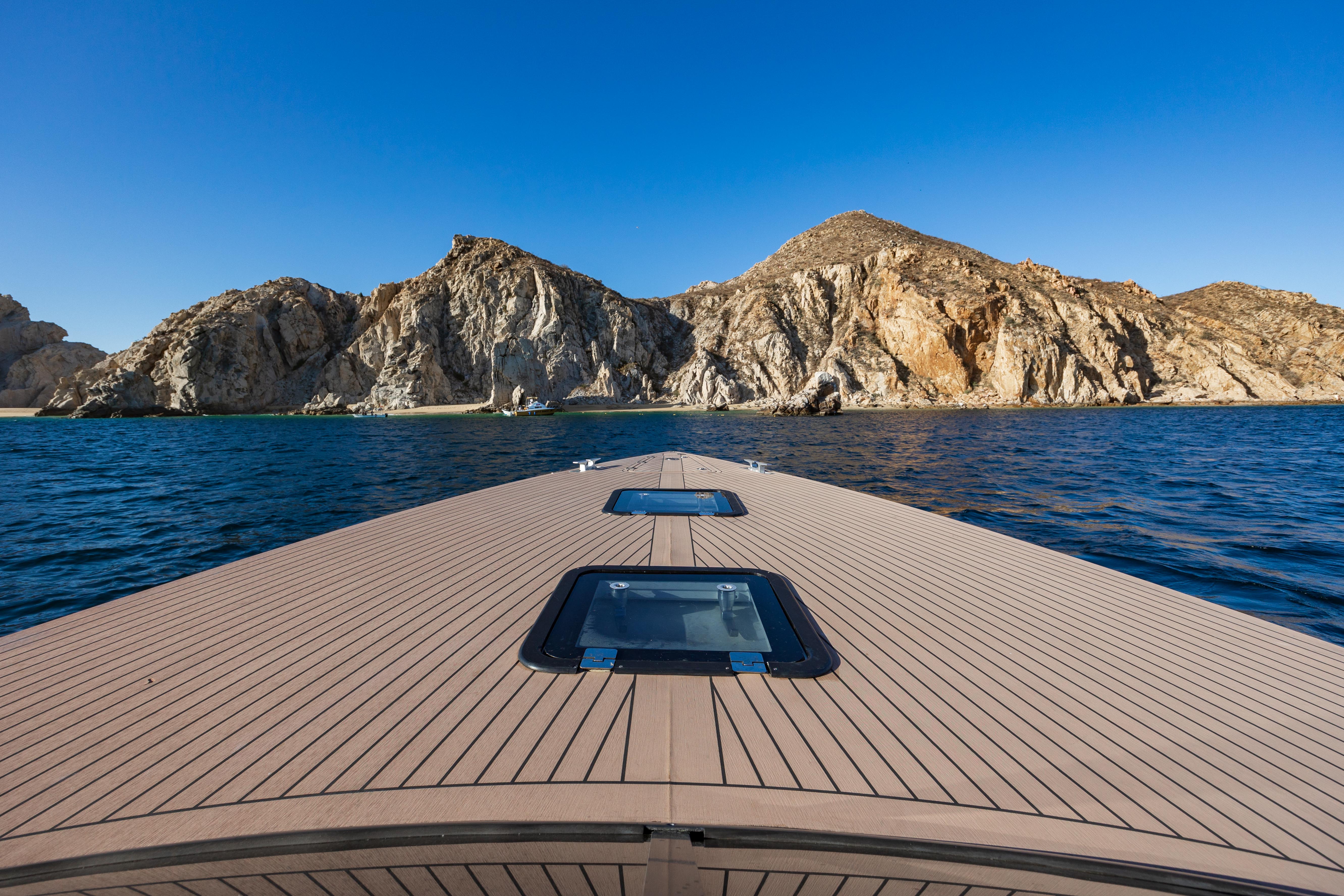 Yacht bow deck pointing toward a rugged rocky coastline and small sandy cove, deep blue ocean waters under a clear sunny sky