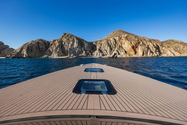 Yacht bow deck pointing toward a rugged rocky coastline and small sandy cove, deep blue ocean waters under a clear sunny sky
