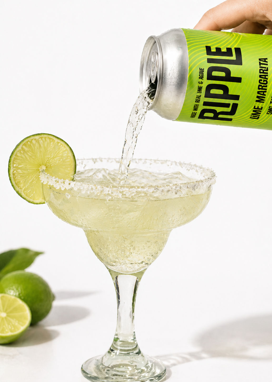 Fizzy canned lime margarita being poured into a salt-rimmed margarita glass, garnished with a lime slice and whole limes in the background