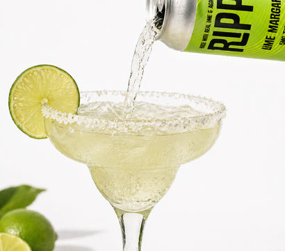 Fizzy canned lime margarita being poured into a salt-rimmed margarita glass, garnished with a lime slice and whole limes in the background
