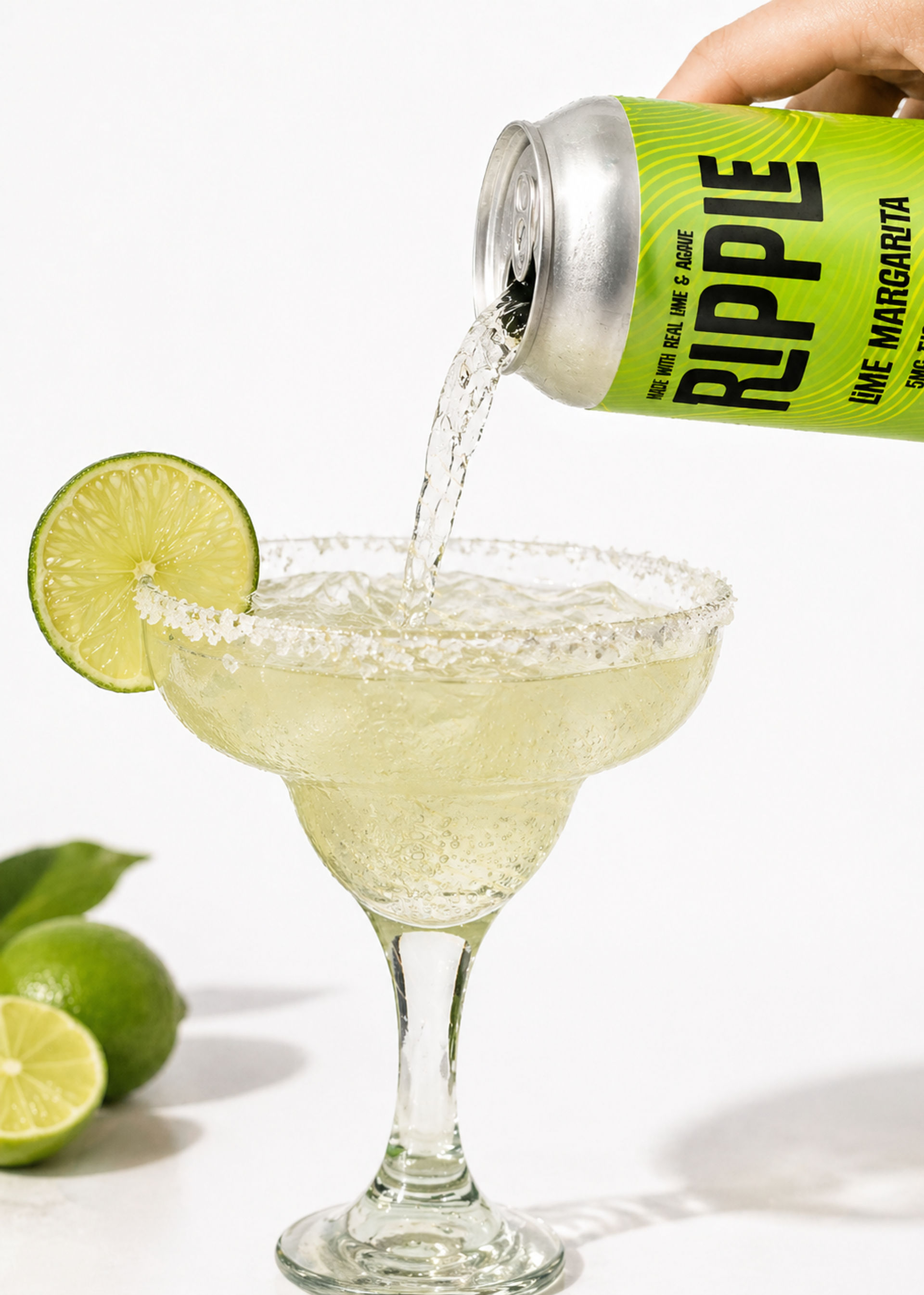 Fizzy canned lime margarita being poured into a salt-rimmed margarita glass, garnished with a lime slice and whole limes in the background