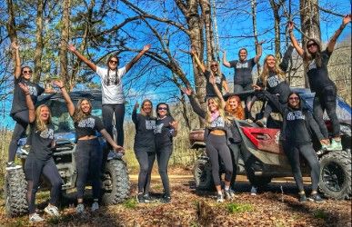 Energetic group of women posing on and around two off‑road side‑by‑side ATVs on a sunny forest trail with bare trees, blue sky, and fallen leaves