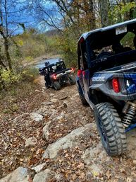 Line of off-road side-by-side UTVs navigating a rocky, leaf-covered trail beside a creek in an autumn woodland