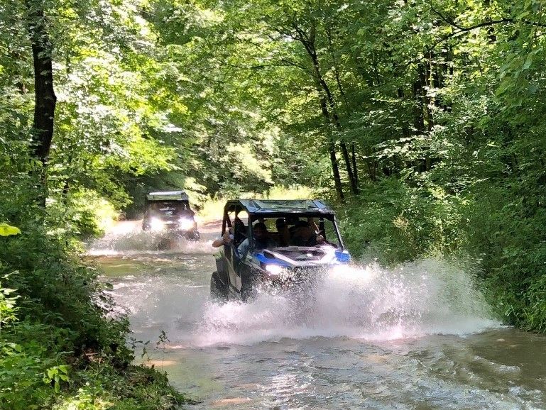 Two off-road UTVs splashing through a shallow water crossing on a sunny, tree-lined forest trail — adventurous summer woodland ride.