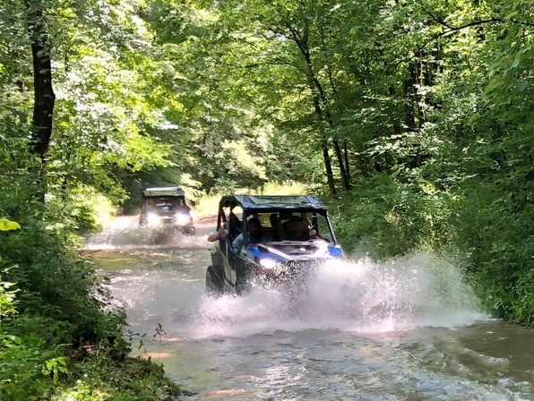 Two off-road UTVs splashing through a shallow water crossing on a sunny, tree-lined forest trail — adventurous summer woodland ride.