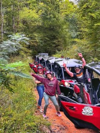 Group of people waving beside red off-road UTVs on a muddy forest trail, enjoying a fun outdoor trail ride through lush green woodland