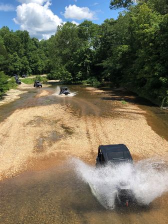 UTVs fording a shallow creek on a sunny summer off-road trail, splashing water across a gravel bar surrounded by dense green forest