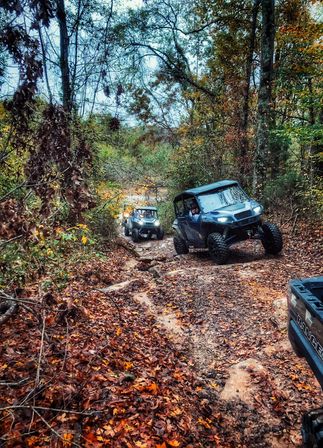 Two off-road UTVs climbing a muddy, rocky forest trail amid colorful autumn foliage and a carpet of fallen leaves.