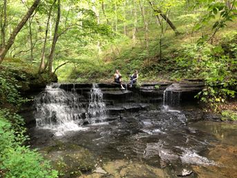 Two hikers perched on a tiered rock ledge above a small cascading woodland waterfall and shallow forest stream, surrounded by lush green trees and mossy rocks.