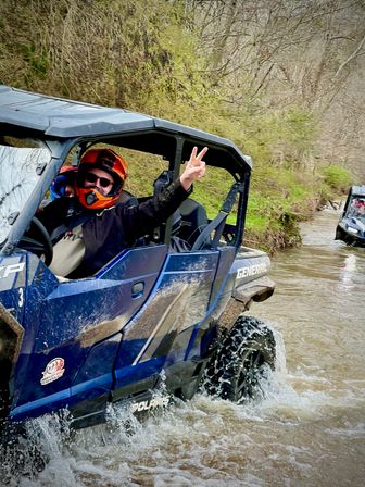 Blue side-by-side UTV splashing through a muddy creek on a wooded off-road trail, driver in an orange helmet flashing a peace sign.