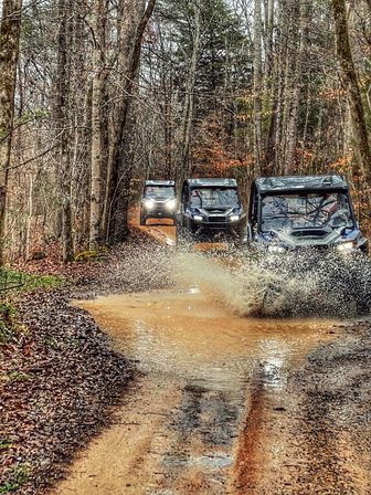 Three off-road UTVs driving a muddy forest trail, the lead vehicle sending a dramatic splash through a large puddle amid bare trees and leaf-covered banks.