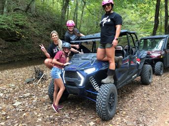 Four friends wearing helmets pose playfully on and beside a blue off-road side-by-side UTV beside a shallow creek on a leafy forest trail — outdoor off-road adventure vibe.