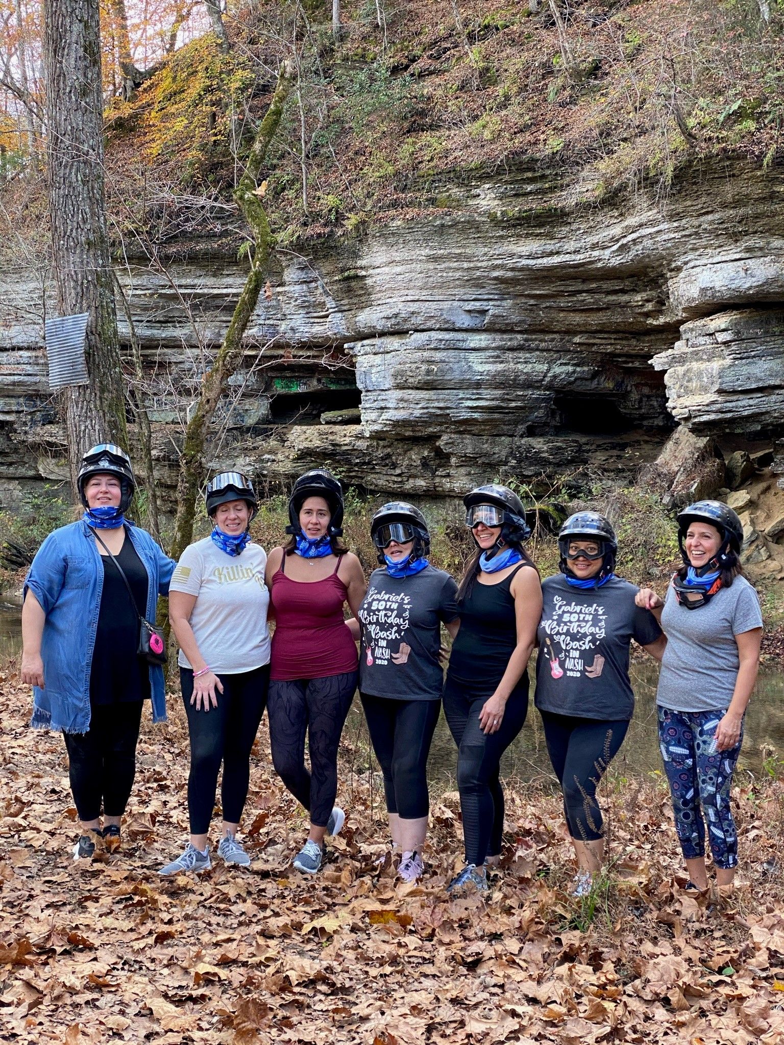 Seven women wearing helmets and blue neck gaiters smiling on a leaf-strewn trail beside layered limestone bluffs and a shallow creek in an autumn forest
