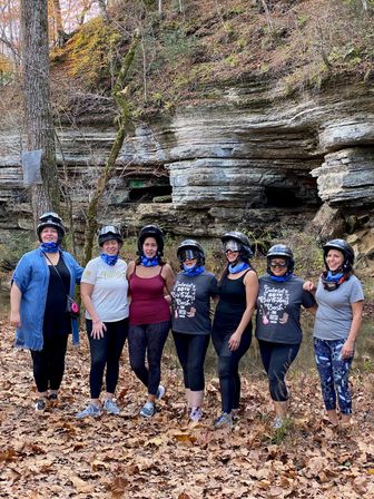 Seven women wearing helmets and blue neck gaiters smiling on a leaf-strewn trail beside layered limestone bluffs and a shallow creek in an autumn forest