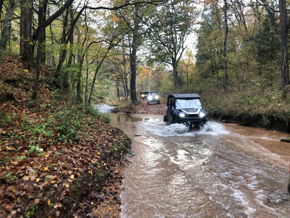 Two off-road UTVs fording a shallow creek on a forest trail amid colorful autumn leaves, headlights on and muddy water splashing