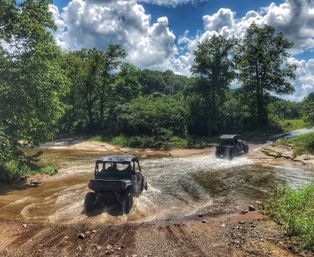 Two off-road UTVs fording a shallow creek on a forest trail, splashing through water under lush green trees and dramatic cumulus clouds on a sunny summer day.