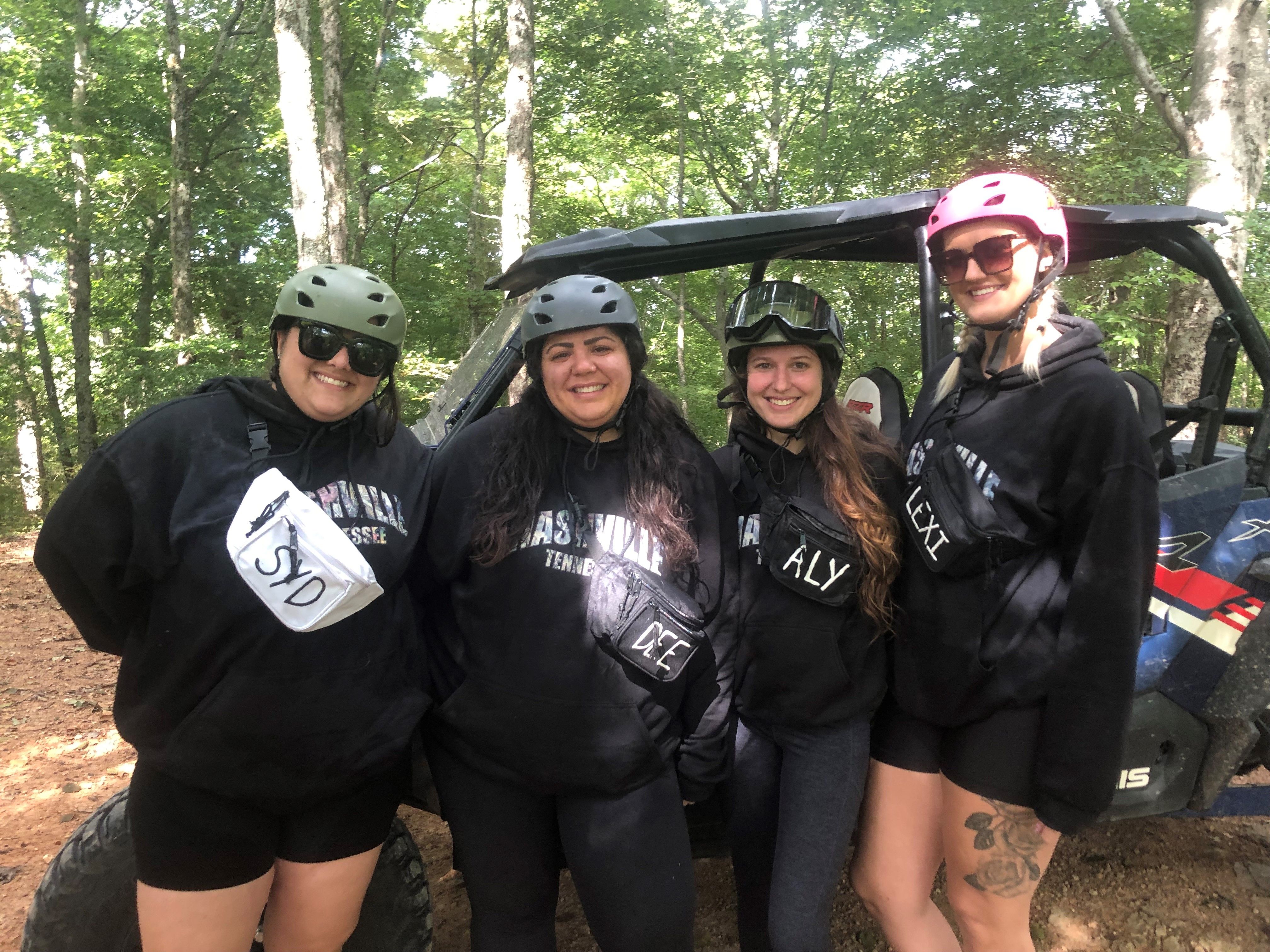 Four friends wearing helmets and black hoodies pose smiling next to a side-by-side UTV on a shaded wooded trail, ready for an off‑road adventure.