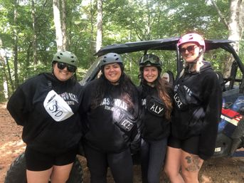 Four friends wearing helmets and black hoodies pose smiling next to a side-by-side UTV on a shaded wooded trail, ready for an off‑road adventure.